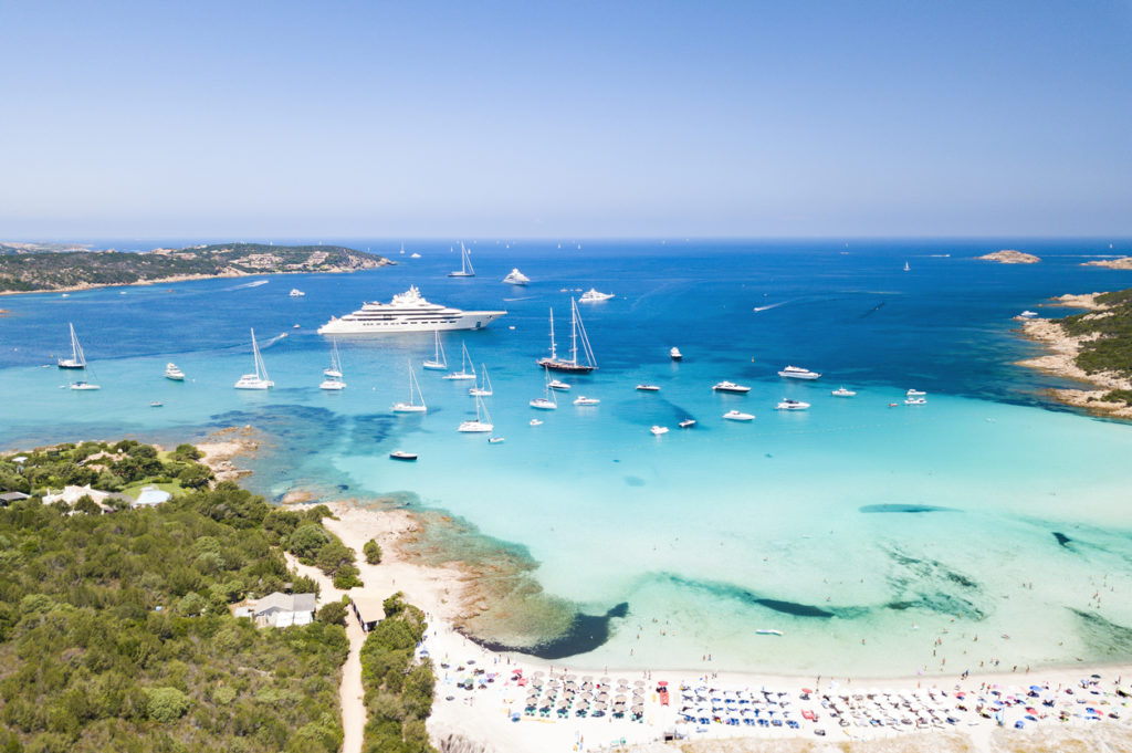 Aerial view of an emerald and transparent Mediterranean sea with a white beach full of beach umbrellas and tourists who relax and take a bath. Gulf of the Great Pevero, Costa Smeralda, Sardinia, Italy.